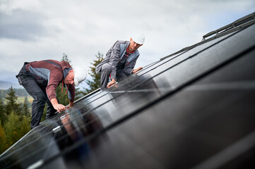 Builders installing photovoltaic solar panels on roof of house. Men engineers in helmets building solar module system with help of hex key. Concept of alternative, renewable energy.