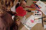 Fototapeta Na drzwi - High angle of hands of child cutting out red paper heart over table with crayons, handmade postcards, watercolors and highlighters