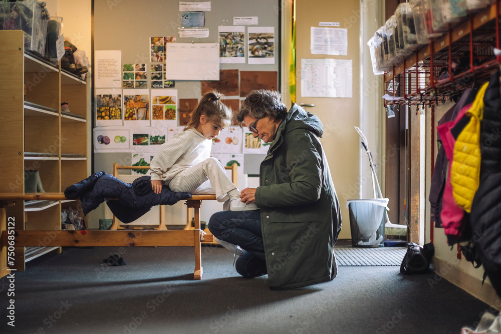 Side view of female teacher helping girl while tying shoe lace in cloakroom at kindergarten