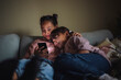 © Maskot - Smiling female siblings sharing smart phone sitting on couch at home