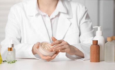  Dermatologist with jar testing cosmetic product at white table indoors, selective focus