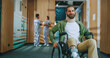 © ihorvsn - Happy smiling young Caucasian patient moving in wheelchair from side to side in hospital is looking at camera and smiling. Successful medical personnel in background. Modern hospital concept.