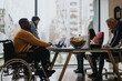 © qunica.com - Inclusive work culture depicted with multi-ethnic team including a person with a disability engaging in a collaborative work session in a bright office.
