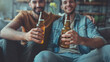 Two hispanic men are sitting on a couch, holding up their beer bottles and smiling at home 