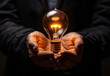 A businessman's hands, holding an idea light bulb on a black background, in light amber and orange tones with eccentric props.