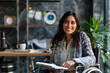 © jex - A successful disability South Asian businesswoman in a wheelchair sits at her desk, exuding warmth and confidence. Holding a notepad and pen, she is poised to take notes, smile directed at the camera