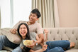 © bongkarn - A cheerful and joyful young Asian couple is watching a sports match or game show together at home.