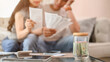 © bongkarn - A jar of coins on a coffee table with a stressed couple planning their finances.