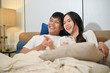 © bongkarn - A happy young Asian couple in pajamas is enjoying watching television or a movie together in bed.
