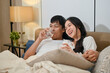 © bongkarn - A happy young Asian couple in pajamas is enjoying watching television or a movie together in bed.