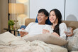 © bongkarn - A happy young Asian couple in pajamas is enjoying watching television or a movie together in bed.