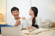 © bongkarn - A cheerful Asian couple is laughing, enjoying watching a movie on a laptop in bed together.