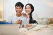 © bongkarn - A cheerful Asian couple is laughing, enjoying watching a movie on a laptop in bed together.
