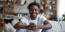 A cheerful young African American man sits indoors, smiling as he uses his smartphone for business.