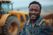 A professional black male engineer in overalls smiles in front of a large yellow tractor against the background of a freshly plowed field, symbolizing progress and productivity.