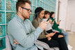 © dikushin - Side view of focused young diverse ethnicity candidates using mobile phones while waiting job interview in modern office lobby. Bored male and female for vacancy sitting on chairs in queue corridor.
