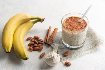 Wall Mural - Glass with protein drink, healthy milkshake smoothie on white table with bananas, protein powder in measuring spoon, almond nuts and cinnamon sticks