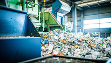 A large pile of unsorted recyclable materials sits inside a warehouse, ready to be processed and sorted at a recycling facility