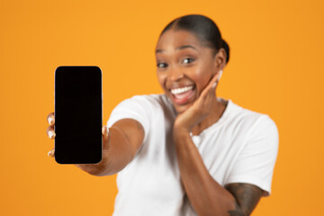Excited black woman extends arm showing smartphone empty screen, studio