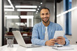 © Tetiana - Portrait of a young African-American man sitting at a desk in a modern office, holding a tablet and smiling at the camera