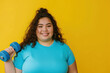 Young happy chubby overweight plus size big fat fit Latin woman wear blue top warm up training hold in hand dumbbell look camera isolated on plain yellow background studio home gym.