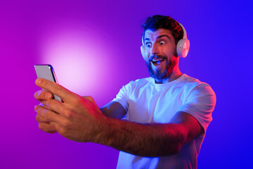 Wall Mural - Surprised Guy Wearing Wireless Headphones Looks At Mobile Phone, Studio