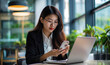 © Jaroonsak - Beautiful Asian business woman sitting at a desk in the office using a laptop and using a mobile phone to search for information online.
