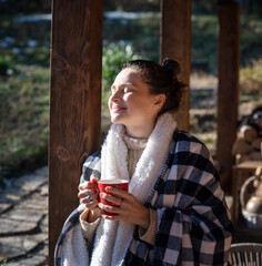 Wall Mural - Young woman enjoying a sunny early spring morning on the terrace, wrapping herself in a blanket and drinking hot drink