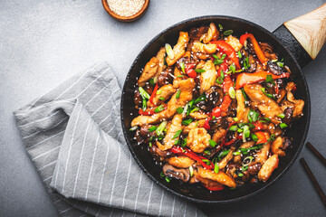 Wall Mural - Asian stir fry chicken  slices with red paprika, mushrooms, chives and sesame seeds in frying pan. Gray kitchen table background, top view