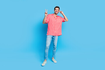 Poster - Full size photo of good mood funky man dressed striped shirt denim pants showing v-sign on eye smiling isolated on blue color background