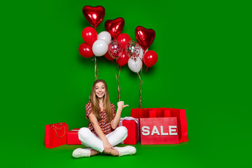 Sticker - Full size photo of cheerful adorable girl sit near gifts indicating at sale shopping bags near balloons isolated on green color background