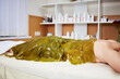 Woman lies on couch with her body algae packed in beauty salon.