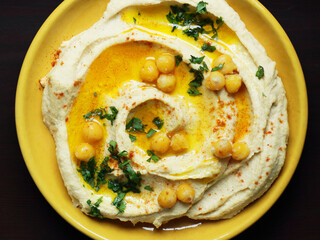 Wall Mural - A plate with hummus with oil, parsley and red pepper along wirh ingredients on wooden board on bright textile, overhead top view, healthy legume food concept