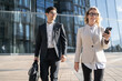 © muse studio - Business professionals in smart attire engaging with technology while walking outside a modern glass building, portraying a dynamic work environment.