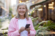 © Liubomir - Portrait of a smiling senior woman in a pink shirt standing outside in the middle of the city and using a mobile phone, looking confidently at the camera.
