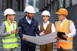 © Liubomir - Focused diverse group of engineers with hard hats discussing blueprints in front of an office building.