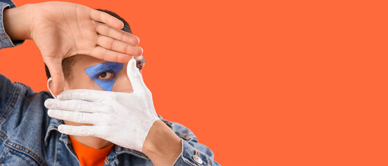 Wall Mural - Young man with painted body making frame gesture on orange background, closeup