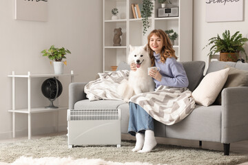 Canvas Print - Young woman with Samoyed dog and tea cup on sofa warming near radiator at home