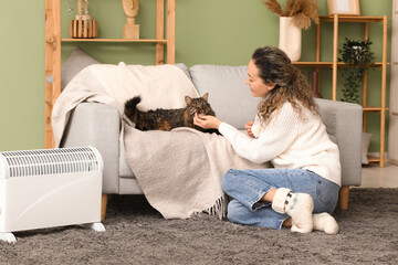 Poster - Happy mature woman petting cute cat on sofa near electric heater at home