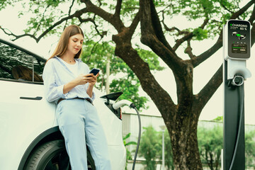 Young woman recharge EV electric vehicle battery from EV charging station and using smartphone online banking to pay for electricity in city park. Eco friendly vehicle travel with EV car. Exalt