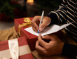 © Mikolette Moller/peopleimages.com - Pen, hands and man with card and gift for Christmas event or party at home for family. Celebration, paper and closeup of male person writing letter with present boxes for xmas festive holiday.