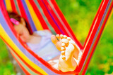 Fototapeta Natura - Young beautiful girl sleeping in a hammock with bare feet, relaxing and enjoying a lovely sunny summer day. Green vegetation in background. Safety and happy childhood concept