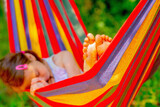 Fototapeta Natura - Young beautiful girl sleeping in a hammock with bare feet, relaxing and enjoying a lovely sunny summer day. Green vegetation in background. Selective focus on bare feet