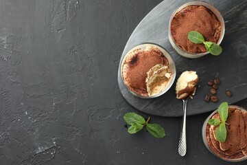 Wall Mural - Delicious tiramisu in glasses, spoon, mint leaves and coffee beans on black table, top view. Space for text