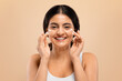 © Prostock-studio - Beauty Routine. Happy young indian woman applying facial cream on her cheeks