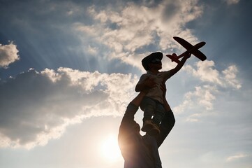 Wall Mural - Silhouettes, sky with clouds, holding toy plane. Father and little son are playing and having fun outdoors