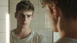 Young man looking at himself in mirror in bathroom. The man has a sad look, depressed mood.