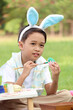 © Stella - Cute Asian boy with bunny ears painting eggs with paintbrush while sitting on green grass meadow in nature garden. Kid celebrating Easter holiday outdoor. Happy child has fun in park on Easter