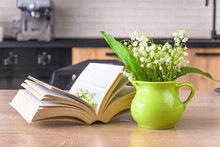 Spring Holidays Concept; Happy May Day Concept; Bouquet Of Lilies Of The Valley In Green Jug, Open Book On A Wooden Table In A Kitchen