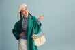 Fashionable happy smiling woman wearing beret, green coat, turtleneck, pearl necklace, holding trendy white leather bag with snowdrops, posing on blue background. Copy, empty, blank space for text